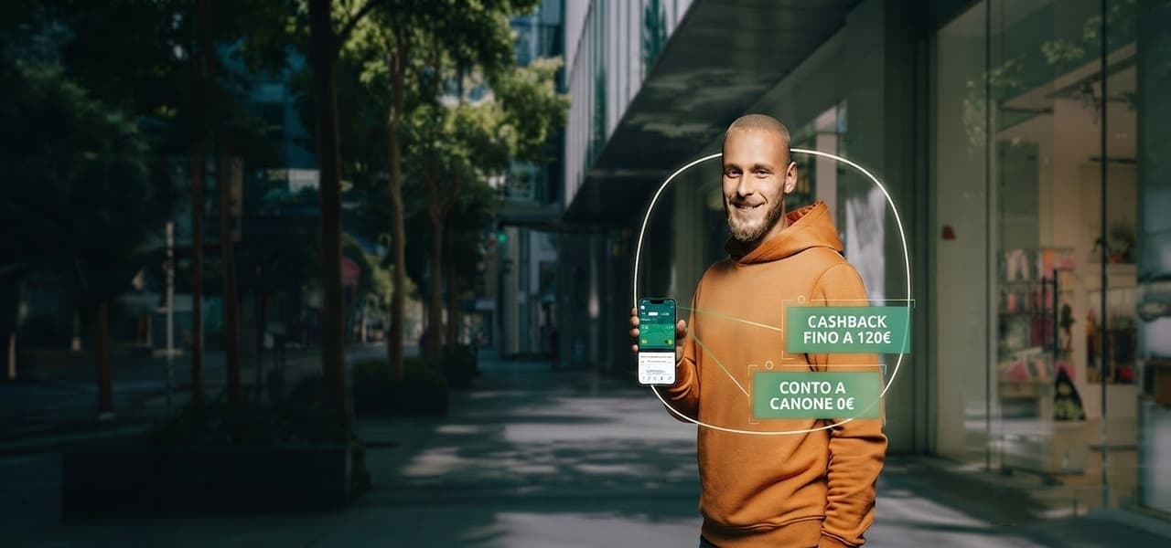 Uomo sorridente in felpa arancione tiene in mano uno smartphone con grafica circolare che segnala cashback fino a 120€ e conto a canone 0€, in una strada cittadina con negozi. | Webranking